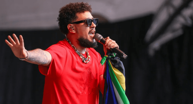 A man wearing sunglasses and a red shirt sings into a microphone on stage, holding a colorful flag, channeling "The World Is Yours" energy with one hand raised.