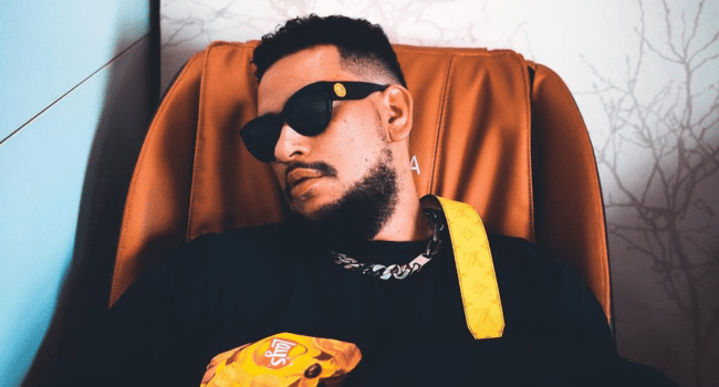 A man with a beard and sunglasses sits in a brown chair, wearing a black shirt and chain necklace, the yellow patterned strap across his chest hinting at Raphael Benza’s special album with AKA Mass Country vibes.