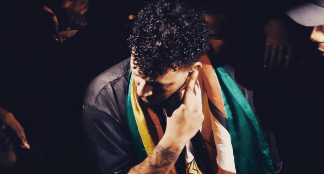 A person with curly hair looks down, draped in a multicolored flag, surrounded by people in dim lighting—a scene reminiscent of Hopewell Chin’ono’s somber moments during his time in Zimbabwean prison.