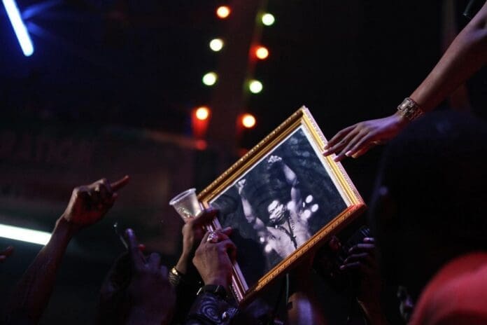 A group of hands reach toward a framed black-and-white photo of Fela Kuti, held up amid colorful lights in a dark, crowded setting that pulses with the energy of African music.