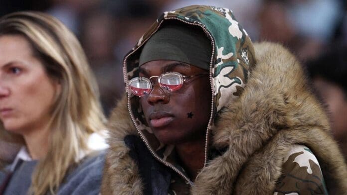 A person wearing a camouflage fur-trimmed jacket, glasses, and a headscarf sits indoors next to another person, discussing the Top 10 African countries shaping today’s music industry.