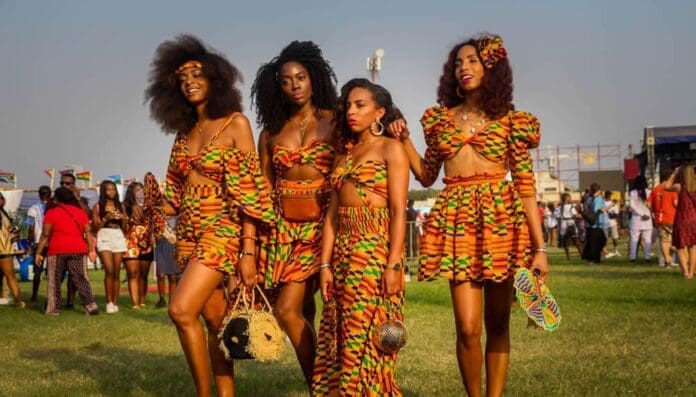 Four women stand together outdoors in matching colorful patterned outfits, embracing the spirit of African music festivals, with other people and event tents visible in the background.