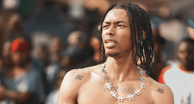 A shirtless man with braided hair and tattoos looks nonchalant as he stands outdoors among a crowd, wearing a large chain necklace.