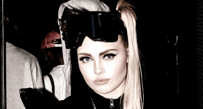 A woman with long blonde hair in a high ponytail, wearing dark goggles on her head, stands in front of a dark background with other people behind her, channeling BABYG!RL ZAE's bold style.