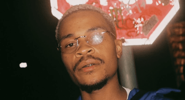 A man with short blond hair and glasses stands outside at night, with a red, graffiti-covered street sign in the background—perhaps waiting for inspiration for Blue Pappi’s new EP, Uthando Olufudumele.