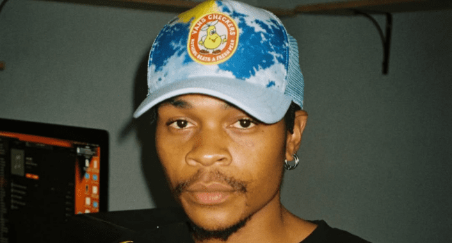 A person wearing a blue and white cap with a "Vato Checkers" logo stands indoors, facing the camera. A computer monitor is visible in the background, hinting at their love for Young Stunna and Imali Entsha.