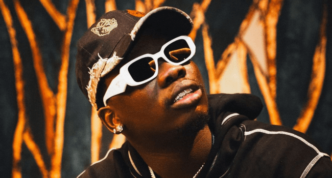 A man, resembling Blxckie, wears white sunglasses, a black cap with patches, and a black hoodie as he looks upward. The background features abstract orange and black patterns.