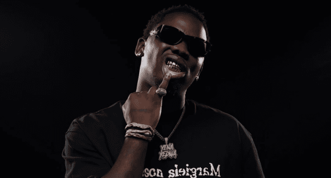 A person wearing sunglasses and jewelry channels Young Stunna as they point to their gold grills while posing against a black background.