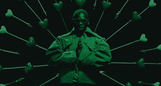 A person in a jacket and sunglasses stands in front of a backdrop featuring heart-shaped arrows pointing outward—a scene reminiscent of Blxckie’s iconic 4LUV2 style.