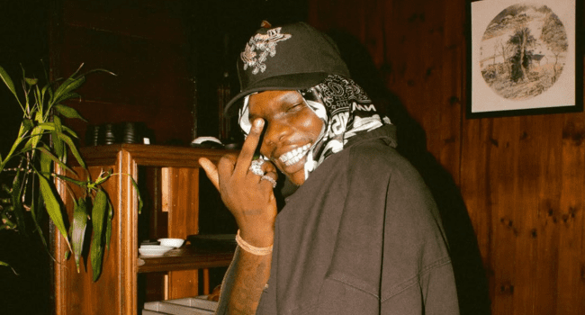 A person wearing a cap and bandana smiles and holds up their middle finger in a dimly lit room with wooden walls, a plant nearby, and a 4LUV2 playlist on Audiomack playing softly in the background.