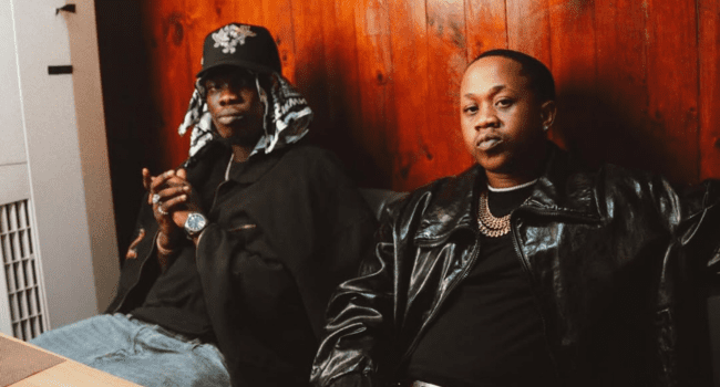 Two men, one resembling Young Stunna with a cap and jewelry, sit side by side in a dimly lit room with wood-paneled walls; both wear dark jackets and gold chains.