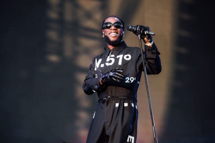 A man wearing sunglasses and black gloves sings into a microphone on stage, dressed in a dark outfit with white numbers and letters printed on it, channeling the bold style of Burna Boy at an NME-featured Afrobeats music event.
