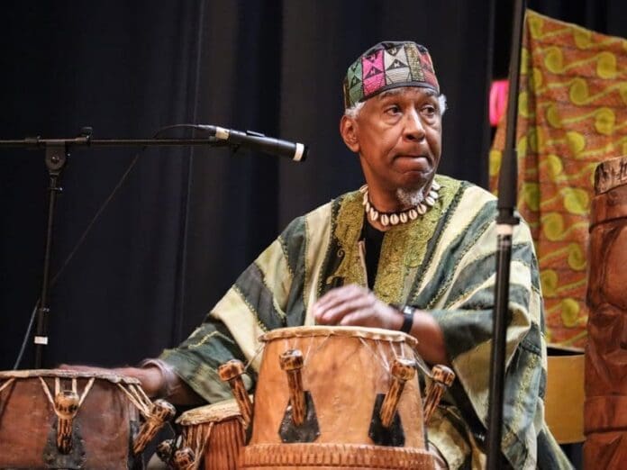 An older man wearing traditional African clothing and a patterned cap plays hand drums on stage, filling the Ukrainian Hall with vibrant African music as a microphone captures every beat.
