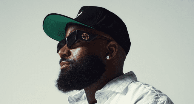A man with a full beard, channeling Cassper Nyovest’s signature style, wears a black cap, dark sunglasses, stud earrings, and a white shirt, looking to the side against a plain background.
