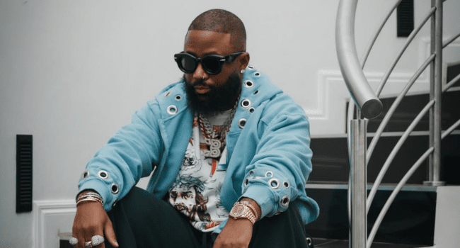 A man with a beard and sunglasses sits on stairs, wearing a blue jacket with silver eyelets, a graphic t-shirt, and several jewelry pieces, channeling cool SA Hip-Hop vibes reminiscent of DJ Maphorisa’s bold street style.