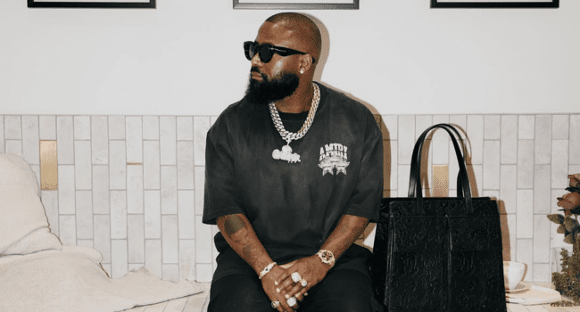 A man with a beard and sunglasses, resembling Emtee, sits on a bed wearing a black T-shirt and jewelry, with a large black bag beside him.