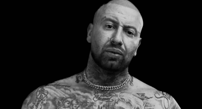 A bald, bearded man with visible tattoos on his chest and neck wears a thick chain necklace and looks directly at the camera against a black background, capturing Chad Da Don’s journey of addiction recovery.