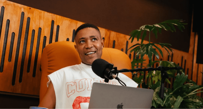 A person sits at a desk with a laptop and microphone, wearing a sleeveless shirt, appearing to discuss artist branding in a podcast studio with plants and wooden paneling in the background—perhaps even interviewing K1llbrady or DJ Speedsta.