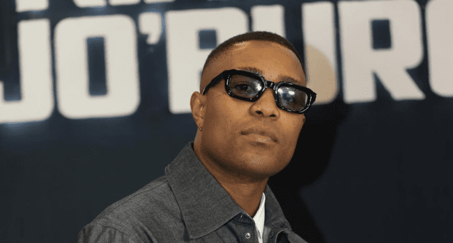 DJ-Speedsta-5.png A person wearing sunglasses and a gray collared shirt poses in front of a dark background with large, blurred white text, capturing the cool vibe of SA Hip-Hop.