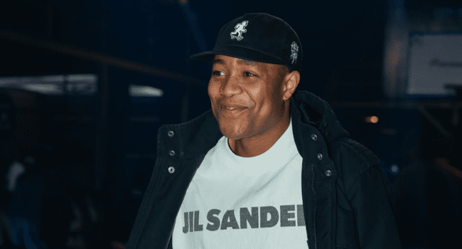 A person wearing a black baseball cap, black jacket, and a white "JIL SANDER" t-shirt is smiling in a dimly lit indoor setting, capturing the cool vibe of The Hip Hop Floor in Soweto.
