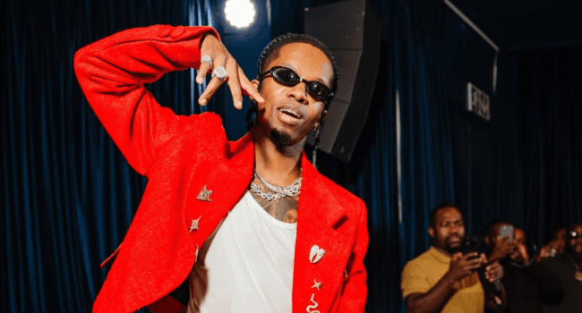 A man wearing a bright red jacket, white shirt, and sunglasses poses with his hand raised, while several people in the background take photos—capturing the vibrant energy seen in Danya Devs' Bayede album and love songs.