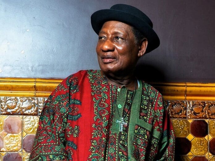 Ghanian musical pioneer Ebo Taylor stands in front of a tiled wall, wearing a patterned red and green traditional outfit and black hat, looking to the side—a portrait capturing his legacy for Far Out Magazine.