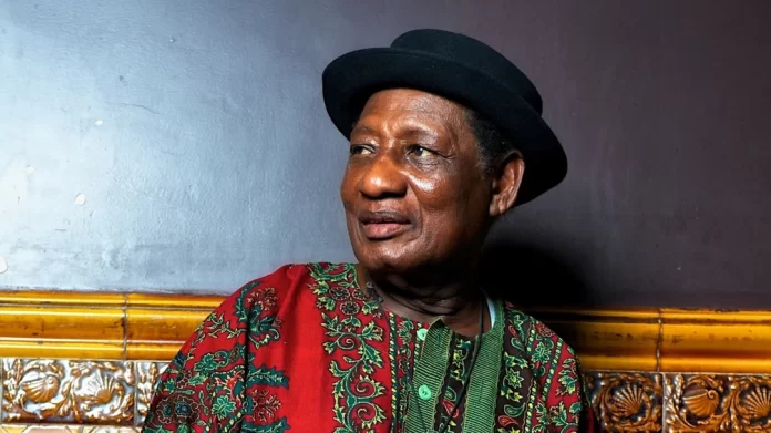 An older man wearing a black hat and a red patterned shirt sits against a dark wall with a decorative yellow border, evoking the timeless style of Ghanaian highlife legend Ebo Taylor.