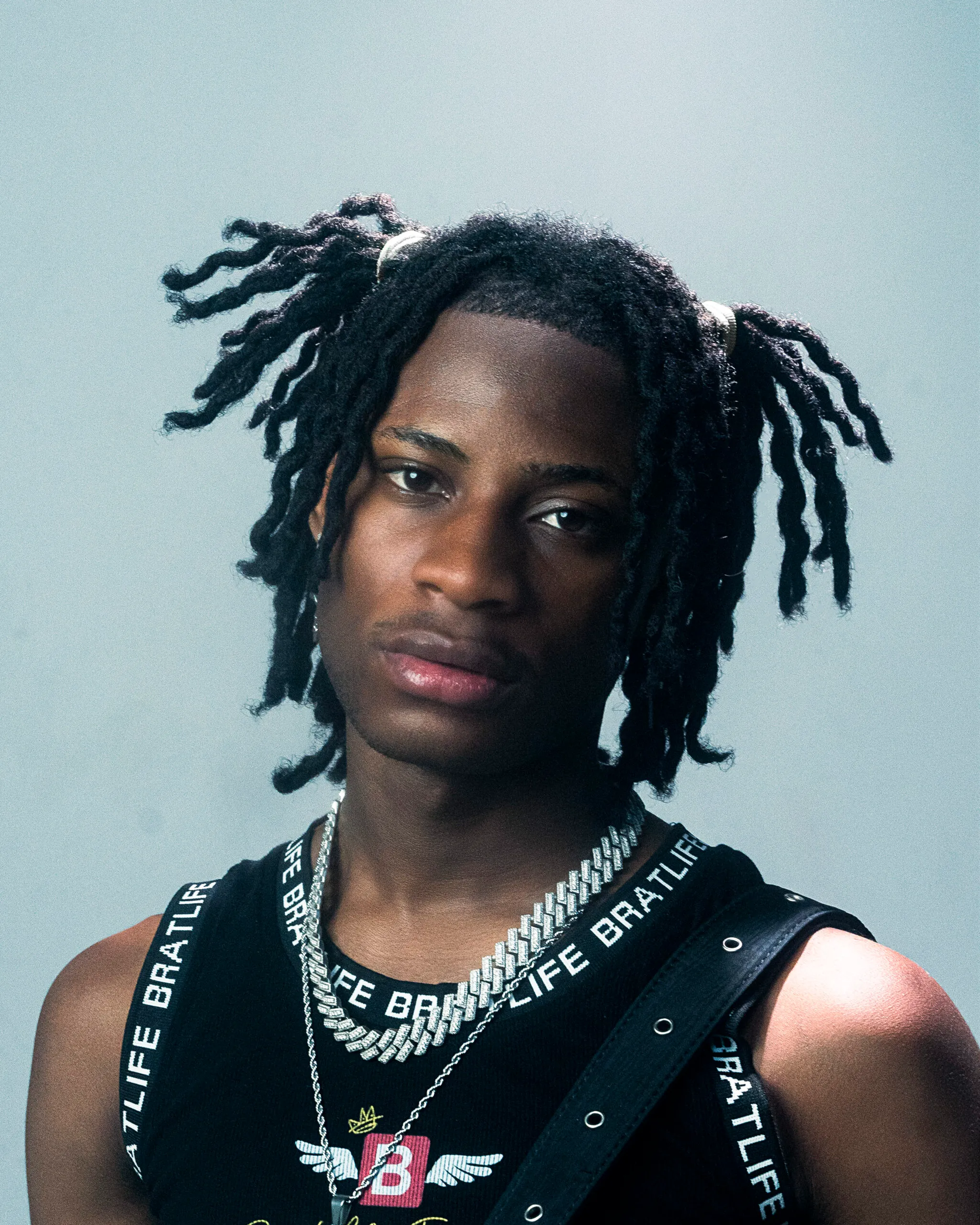 Young person with short, twisted dreadlocks wears a black tank top, two layered chain necklaces, and a serious expression against a plain background, channeling the bold style of Afropop star Khaid.