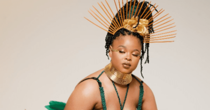 A woman wearing a gold choker necklace, green outfit, and an ornate headpiece with golden spokes poses against a plain background, embodying the vibrant spirit of Afropop and the power to heal hearts through music.