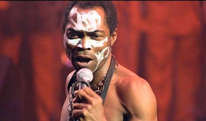 A man with white face paint holds a microphone and sings on stage, wearing a necklace and no shirt, with a red-lit background—capturing the vibrant energy of the Music World.