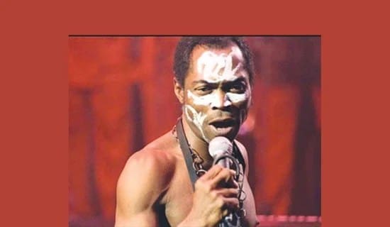 A man with white tribal face paint, inspired by Fela, sings into a microphone, wearing a necklace and no shirt against a reddish-brown background.