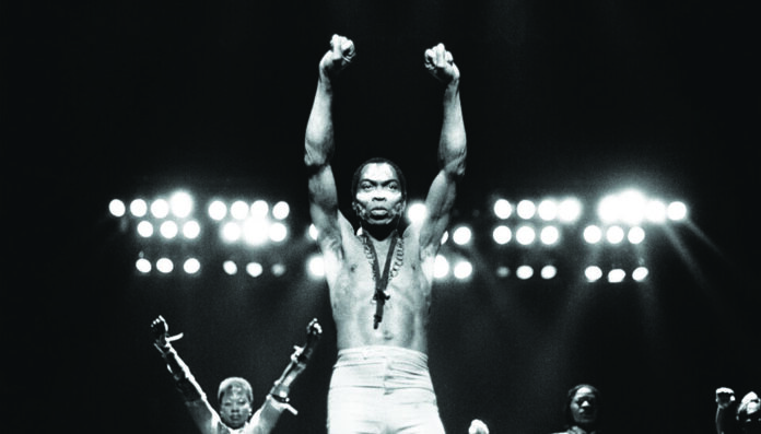 A shirtless man with a necklace stands on stage, fists clenched and arms raised, under bright lights—a powerful scene reminiscent of Fela’s legendary performances; backup performers appear behind him.
