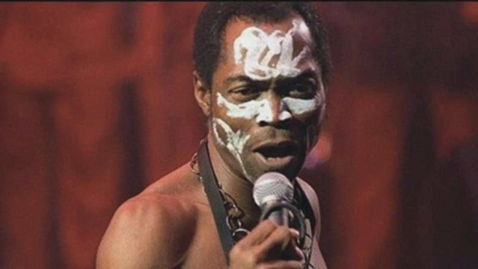 A man with white face paint sings into a microphone on stage in Lagos, wearing no shirt and a beaded necklace. The background is blurred with warm lighting.