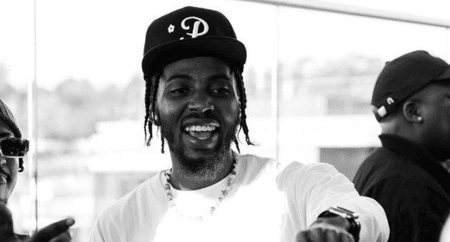 A man wearing a cap and necklace smiles and gestures while standing indoors, embodying the SA Hip-Hop vibe; another person is visible in the background.