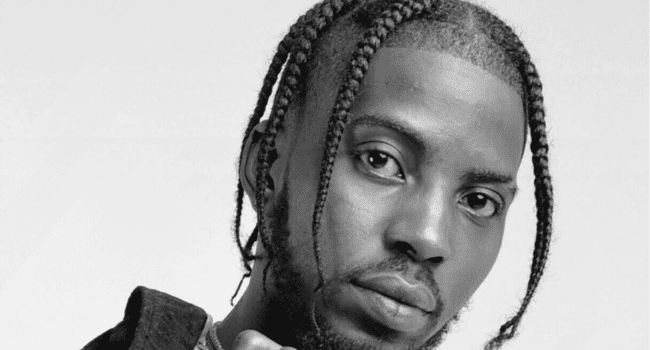 Black and white portrait of Flvme, a man with braided hair, a trimmed beard, and a neutral expression, looking directly at the camera—capturing the essence of his artistry in music production and signature Auto-Tune style.