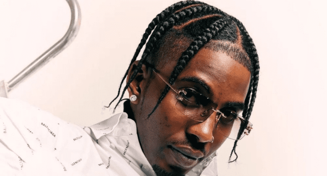 A man with braided hair, wearing glasses, stud earrings, and a white shirt with a subtle pattern, looks towards the camera against a plain background, embodying Flvme's journey to feel free from addiction.