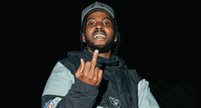 A man wearing a jacket and cap stands against a black background, raising his middle finger toward the camera, channeling the bold energy of Flvme’s BLVCK & WHiTE album.