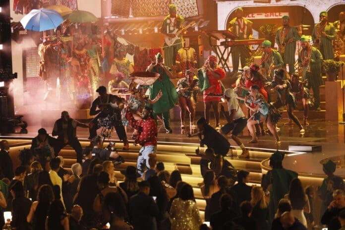 A lively stage performance with brightly costumed dancers and musicians showcases the vibrant rhythms of African music, while some performers interact with the seated audience in front of the stage—a scene reminiscent of Lagos’ dynamic arts culture.