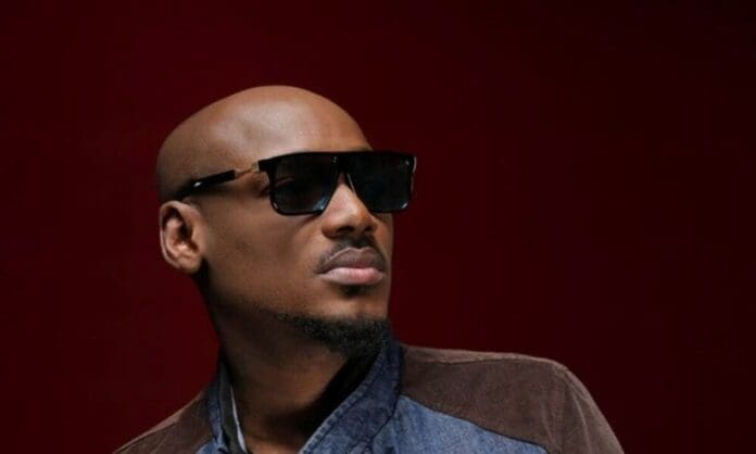 A man wearing dark sunglasses and a denim jacket poses against a dark red background, channeling Afrobeats style.