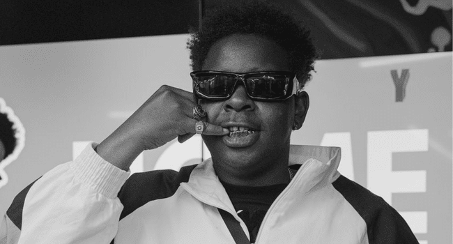 A person wearing sunglasses and a tracksuit jacket makes a phone gesture with their hand and displays a grill on their teeth, channeling Focalistic vibes. The background features partially visible text.