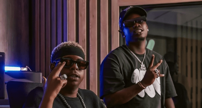 Two people wearing sunglasses and dark clothing pose indoors, both making peace signs with their hands, channeling serious Blxckie hits energy as if they're ready for a top 5 performance of Herc Cut The Lights.