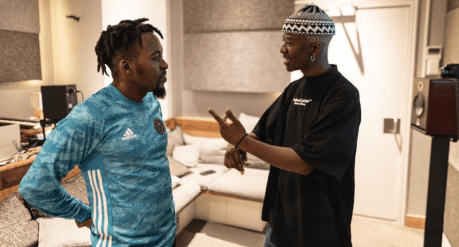 Two men stand in a recording studio having a conversation; one wears a blue sports jersey, the other a black shirt and patterned hat—an everyday scene from Khayelitsha’s vibrant music community.