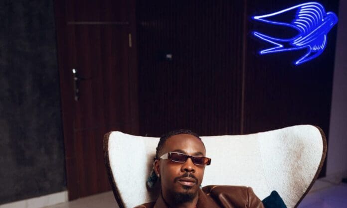 A man wearing sunglasses and a brown jacket sits in a white chair indoors, with a blue neon bird light on the wall behind him, exuding the cool vibe of Afropop singer Young Jonn.