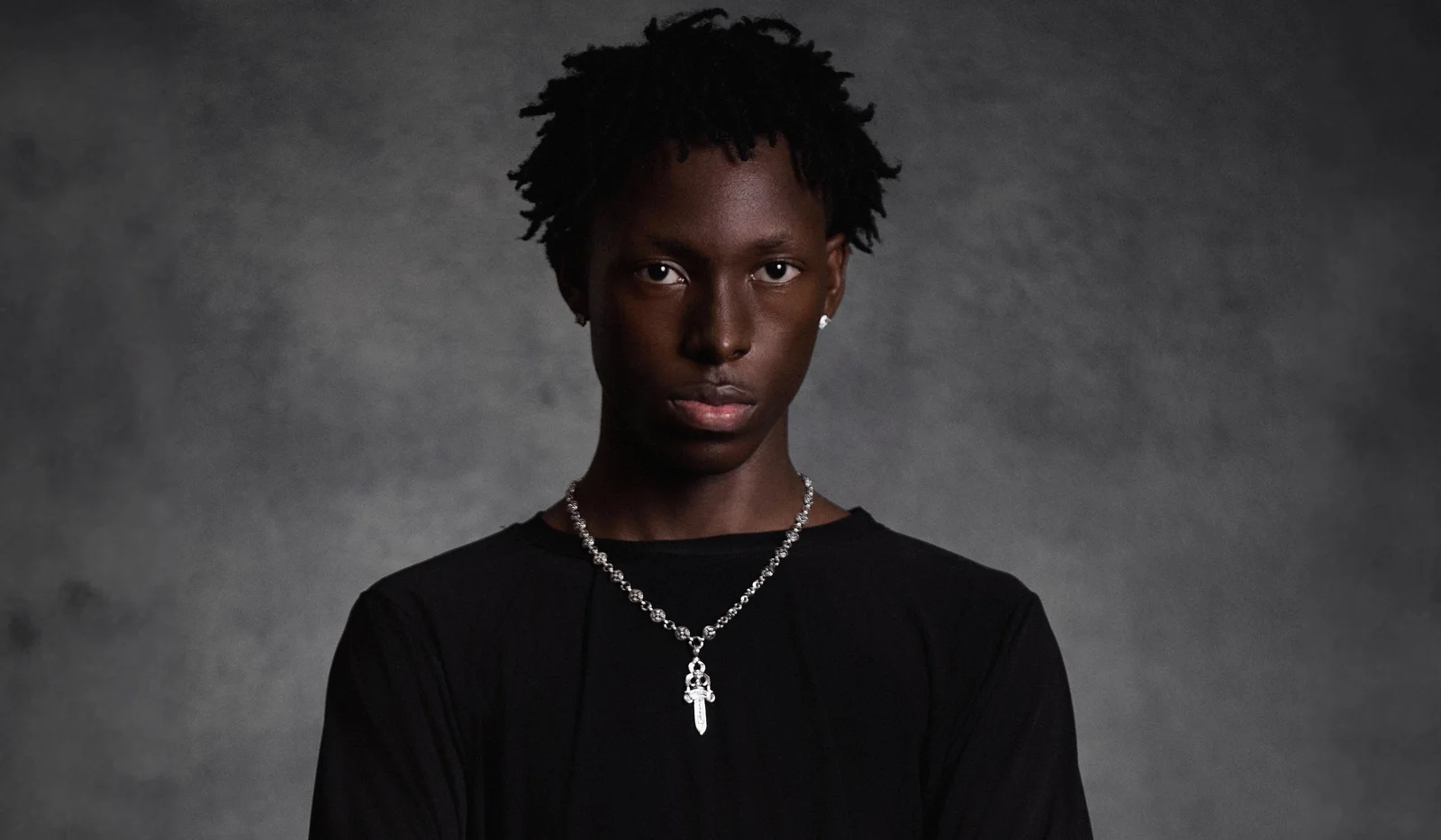 A person with short, textured hair wearing a black shirt and a silver chain necklace stands against a gray textured background, channeling confident Dxtiny energy while looking directly at the camera.