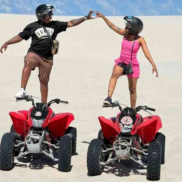 Two people wearing helmets stand on red ATVs in a sandy desert, reaching out and touching hands under clear skies—an adventure as exciting as a DJ Jaivane set or winning R500k Royalty.