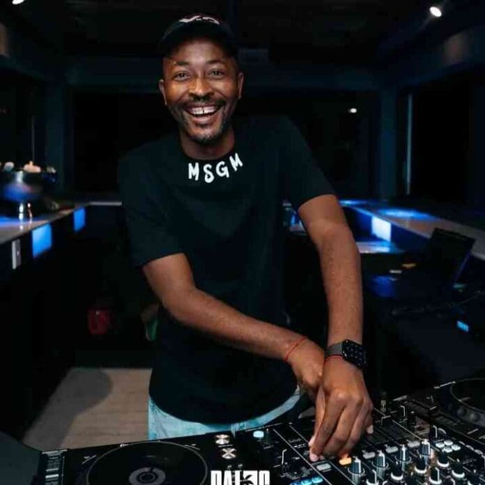 A smiling DJ wearing a black MSGM shirt stands behind a mixing console with one hand on the controls in a dimly lit room, spinning tracks from the Turbang Amapiano Mix for Bandros 2026.