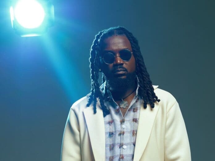 A man with long dreadlocks, wearing round sunglasses, a patterned shirt, and a light-colored blazer stands in front of a bright studio light—channeling Samini’s bold vibe ahead of his eighth album, ORIGIN8A.