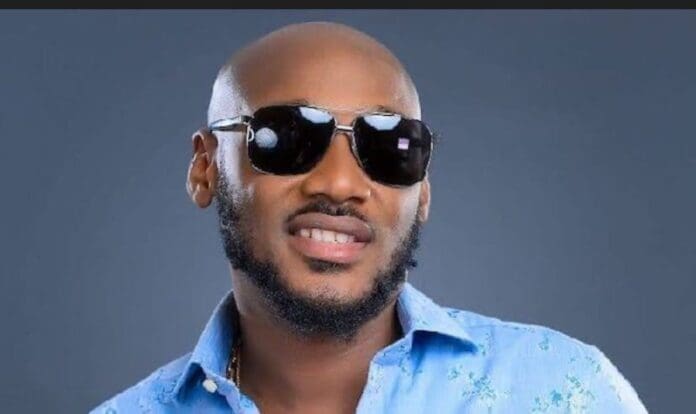 A man wearing a light blue patterned shirt and dark sunglasses poses in front of a plain grey background, channeling the effortless style of 2Face from his iconic Billboard Afrobeats hit, "African Queen.