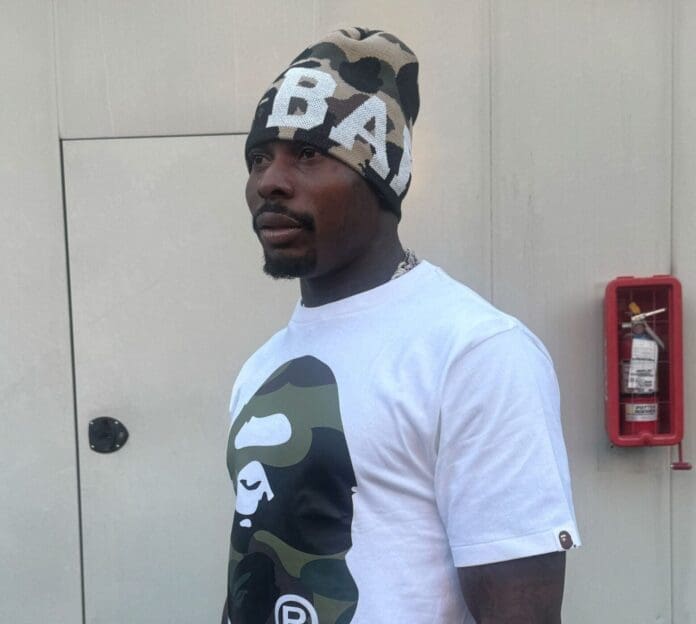 A man wearing a camo beanie and a white BAPE T-shirt stands in front of a gray wall with a red fire extinguisher, channeling an Asake vibe fit for Interview Magazine.