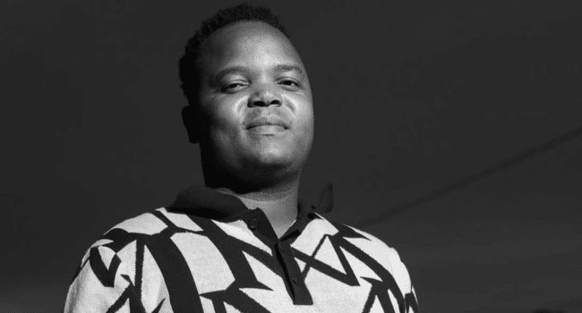 A person stands outdoors, facing forward and wearing a patterned sweater with a collared shirt—a striking black and white photo that echoes the style of Intaba Yase Dubai's new single, Angimbonge Umdali.
